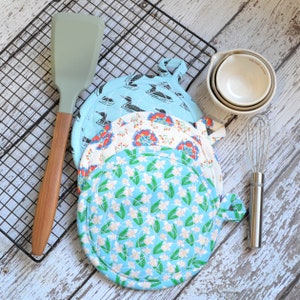 May include: Four quilted pot holders with floral patterns in shades of blue, red, and white. The pot holders have a round shape with a loop handle. The pot holders are stacked on top of each other on a white wooden surface.