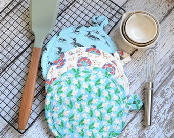 May include: Four quilted pot holders with floral patterns in shades of blue, red, and white. The pot holders have a round shape with a loop handle. The pot holders are stacked on top of each other on a white wooden surface.