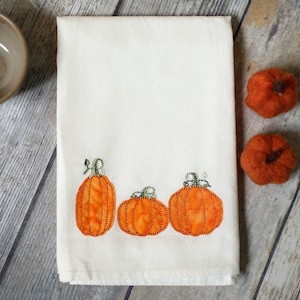 May include: White kitchen towel with three embroidered orange pumpkins with green stems.