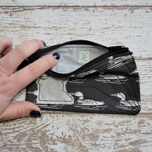 May include: A black zippered pouch with a white outline of the state of Minnesota and a repeating pattern of white loon birds on a black background. The pouch is open and the inside is lined with black fabric. A small white tag with the text "Sew Much More Store" is visible inside the pouch.