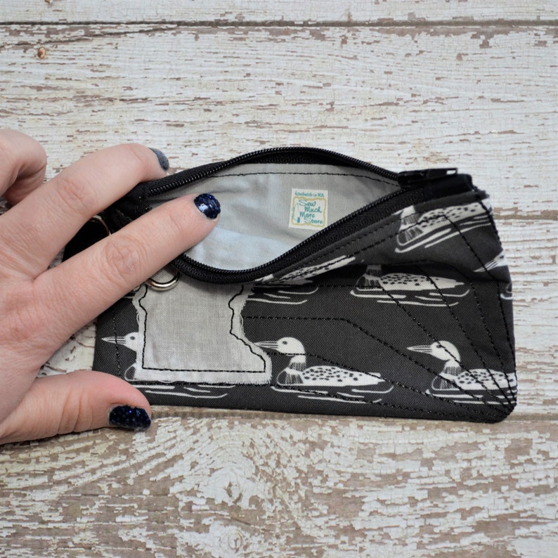 May include: A black zippered pouch with a white outline of the state of Minnesota and a repeating pattern of white loon birds on a black background. The pouch is open and the inside is lined with black fabric. A small white tag with the text "Sew Much More Store" is visible inside the pouch.