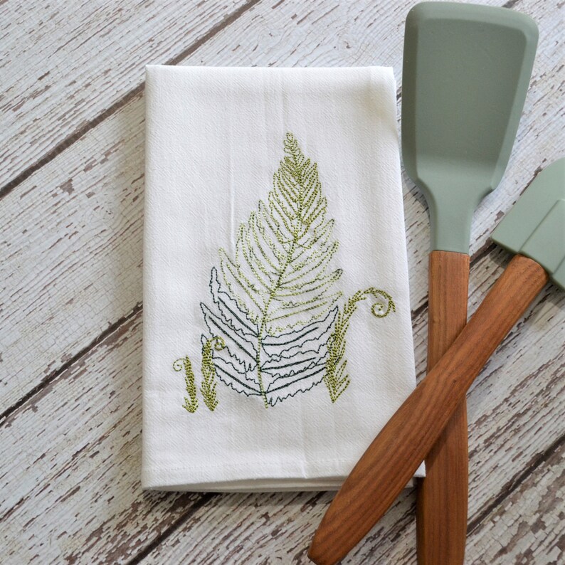 May include: White kitchen towel with an embroidered green fern design. The towel is folded and lying on a wooden surface with two green silicone spatulas with wooden handles.