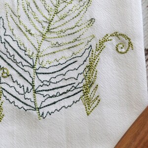 May include: Embroidered design of a green fern leaf with a detailed outline in black thread. The fern is stitched on a white fabric background.
