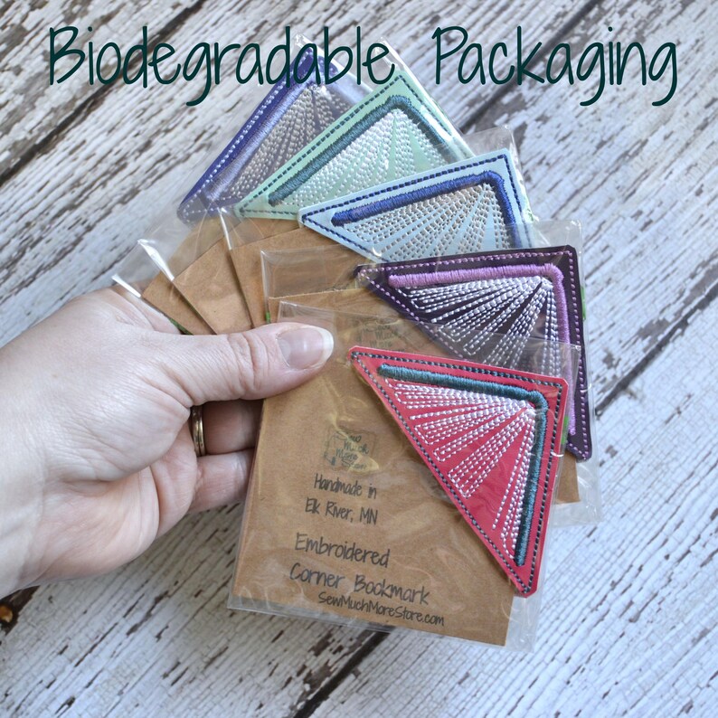 May include: A set of five embroidered corner bookmarks in various colors, packaged in clear plastic bags. The bookmarks are handmade in Elk River, MN and are described as "Embroidered Corner Bookmark" with the website SewMuchMoreStore.com.