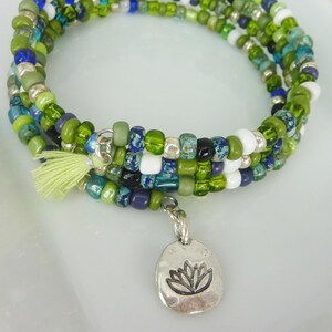 Könnte beinhalten: Wickelarmband mit mehreren Strängen aus grünen, blauen, weißen und silbernen Perlen. Es hat eine kleine, hellgrüne Quaste und einen silbernen Anhänger mit einem Lotusblumen-Design. Das Armband wird auf einer weißen Oberfläche präsentiert.