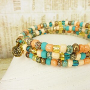 Könnte beinhalten: Mehrsträngiges Wickelarmband mit bunten Perlen in Orange, Türkis, Gold und Creme. Das Armband hat einen kleinen, dekorativen Anhänger und wird auf einer weißen, strukturierten Oberfläche präsentiert.