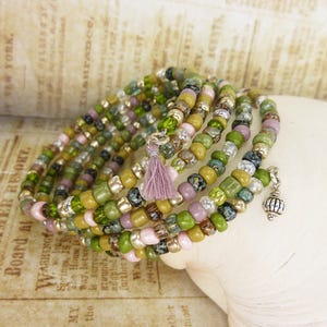 Könnte beinhalten: Mehrsträngiges Wickelarmband mit bunten Perlen in Grün, Rosa und Gold. Es hat eine kleine Lavendelquaste und einen silbernen Anhänger. Die Perlen sind auf einem flexiblen Draht aufgefädelt. Ein stilvolles Accessoire.