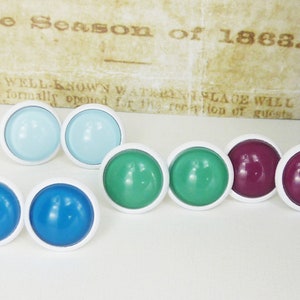 Könnte beinhalten: Eine Sammlung farbenfroher runder Schrankknöpfe mit weißen Rändern. Die Knöpfe sind in Blau-, Grün- und Lilatönen erhältlich. Der Hintergrund enthält den Text "The Season of 1863."