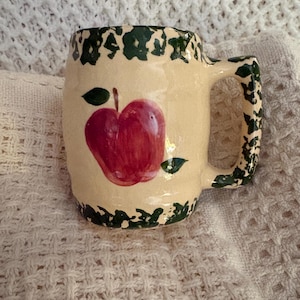 May include: Cream-colored ceramic mug with a red apple design. The mug features a green rim and handle detailing. The apple is painted with a glossy finish, and the mug has a rustic, handmade appearance.