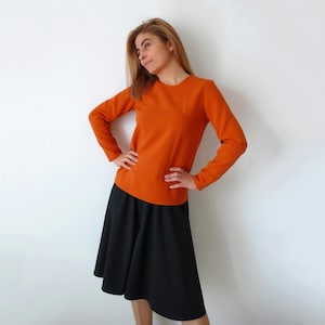May include: A woman wearing a long-sleeved, burnt orange sweater and a black, A-line skirt.