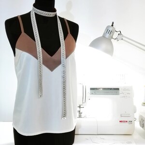 May include: A white tank top with a brown satin trim and a measuring tape draped around the neck. The tank top is on a dress form. A white sewing machine with the text "NECCHI 308" is in the background.