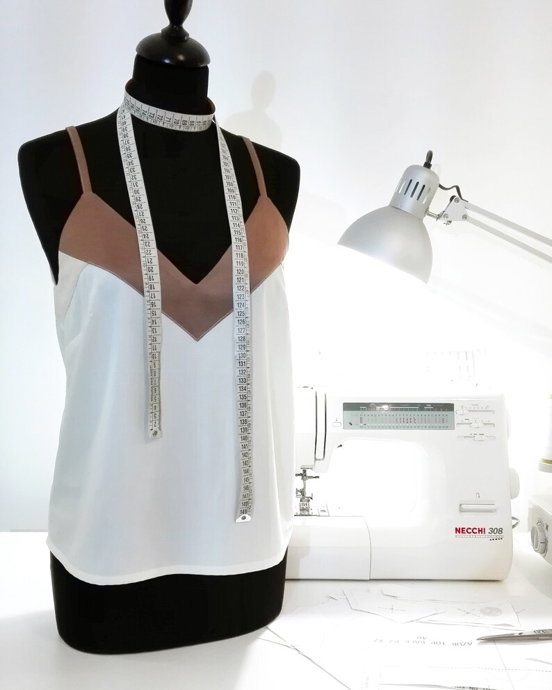 May include: A white tank top with a brown satin trim and a measuring tape draped around the neck. The tank top is on a dress form. A white sewing machine with the text "NECCHI 308" is in the background.