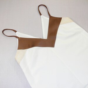 May include: A white sleeveless top with a brown leather trim around the neckline. The top has a V-neck and a relaxed fit.