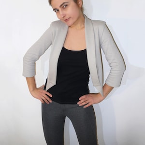 May include: A woman wearing a light grey cropped blazer over a black tank top and grey trousers.
