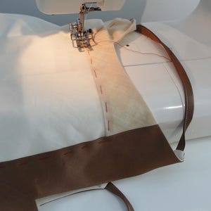 May include: A sewing machine is stitching a brown leather strap to a white fabric. The strap is folded over and the stitching is visible.