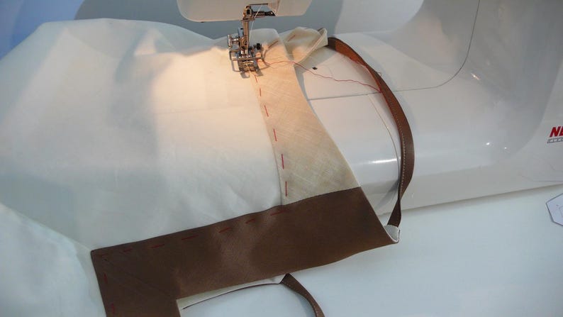 May include: A sewing machine is stitching a brown leather strap to a white fabric. The strap is folded over and the stitching is visible.