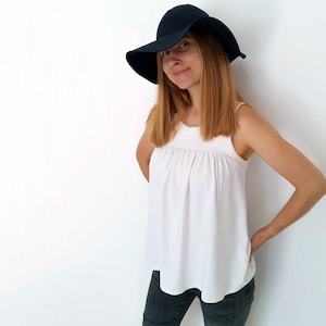 A photo of a smiling woman with long dark blonde hair wearing a white tank-like jersey top with front gathers through the yoke, dark jeans, and a dark blue wide-brimmed hat.