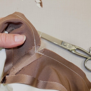 May include: A close-up of a brown fabric being cut with silver scissors. The fabric has a white seam along the edge.