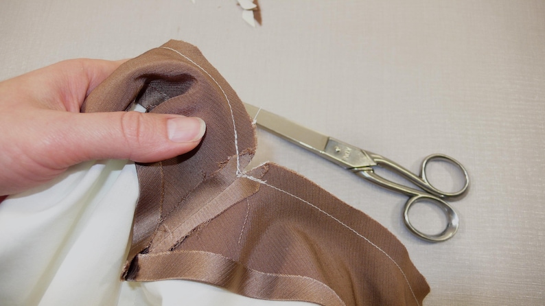 May include: A close-up of a brown fabric being cut with silver scissors. The fabric has a white seam along the edge.