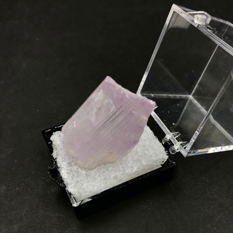 May include: A pale lavender crystal with striations, displayed in a clear plastic box with a black base.