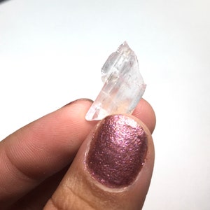 Puede incluir: Un cristal transparente y transl&uacute;cido con una punta puntiaguda y una base plana. El cristal se sostiene en una mano con esmalte de u&ntilde;as rosa.