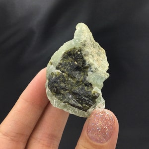 May include: A green and black crystal with a rough, irregular shape. The crystal has a light green color with dark green and black inclusions.