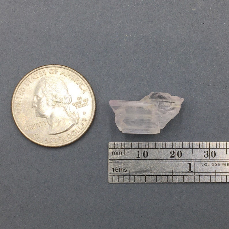 Puede incluir: Un cristal alargado transl&uacute;cido de color rosa p&aacute;lido con una forma ligeramente curvada. El cristal mide aproximadamente 2 cm de largo y est&aacute; sobre una regla con marcas en mil&iacute;metros.