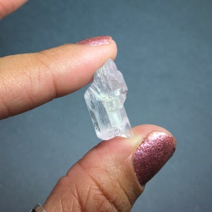 Puede incluir: Un cristal transparente y transl&uacute;cido con un tono rosado, con forma de prisma. El cristal se sostiene en una mano.