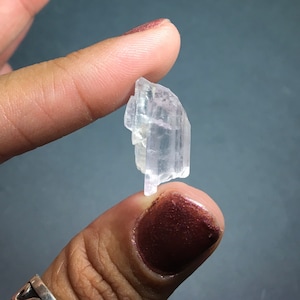 Puede incluir: Un cristal transparente y transl&uacute;cido con un tono rosado. El cristal tiene forma de prisma y tiene m&uacute;ltiples facetas. Se est&aacute; sosteniendo entre dos dedos.