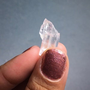 Puede incluir: Un cristal transparente y transl&uacute;cido con un tono rosado, con forma de astilla o punta, sostenido en una mano con esmalte de u&ntilde;as rojo oscuro.