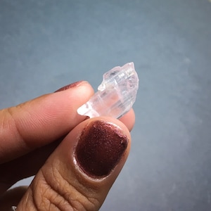Puede incluir: Un cristal transparente y transl&uacute;cido con una punta puntiaguda y estr&iacute;as en su superficie. El cristal se sostiene en una mano con esmalte de u&ntilde;as marr&oacute;n oscuro.