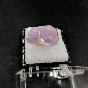 May include: A small, purple, translucent crystal with a white, opaque center. The crystal is sitting on a white, fluffy surface inside a clear plastic box.