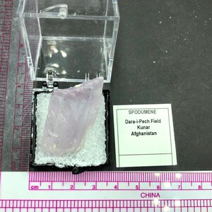 May include: A pink, translucent, rough-cut crystal of spodumene in a small, clear plastic display case. The label reads "SPODUMENE Dara-i-Pech Field Kunar Afghanistan".