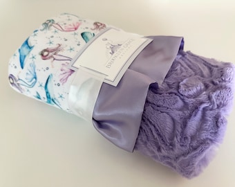 May include: A purple and white baby blanket with a mermaid print on one side and a soft, plush texture on the other. The blanket is folded and tied with a white satin ribbon. The label reads "Brooklyn & Grace Designs".