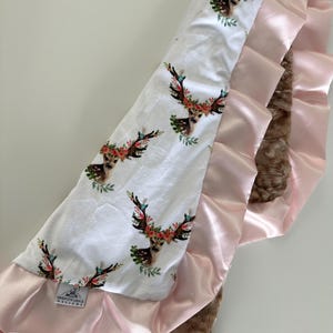 May include: A white baby blanket with a floral deer print on one side and a brown faux fur and pink satin ruffle on the other side. The blanket has a tag that says "Brideson Grace Designs".