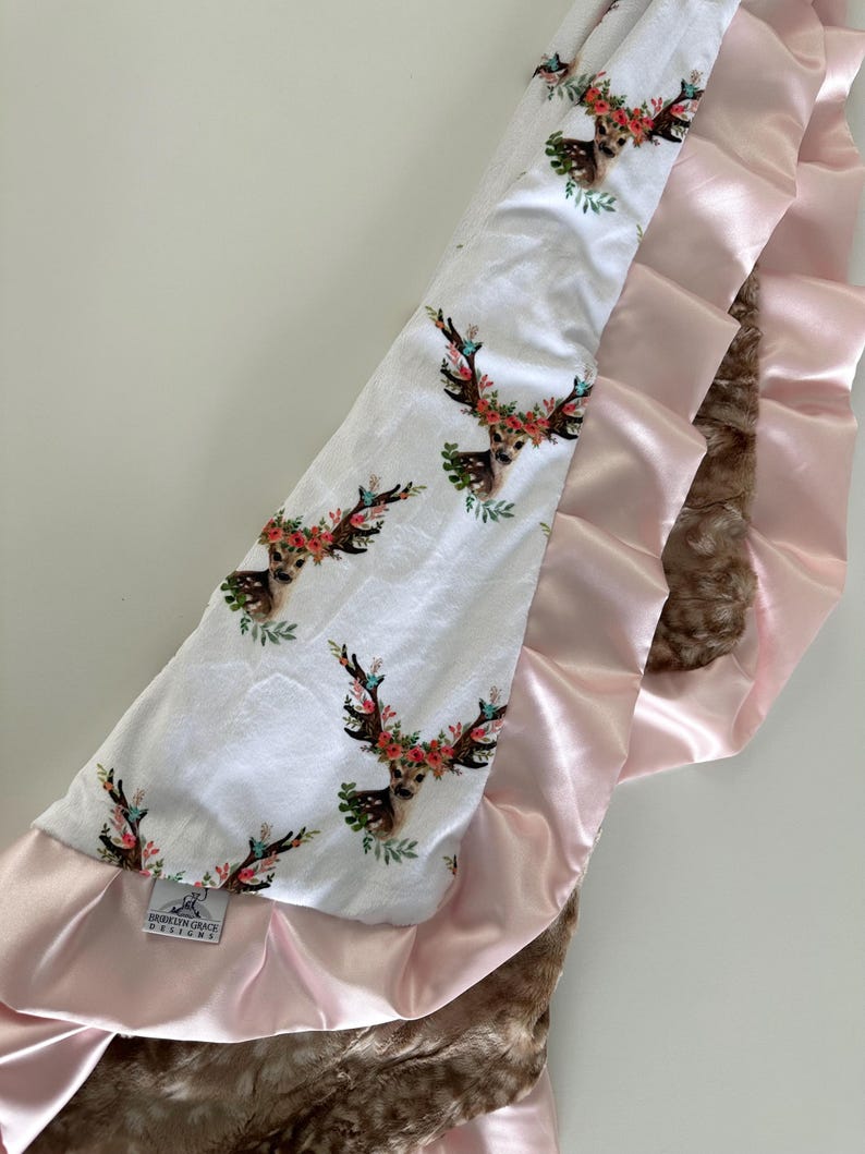 May include: A white baby blanket with a floral deer print on one side and a brown faux fur and pink satin ruffle on the other side. The blanket has a tag that says "Brideson Grace Designs".