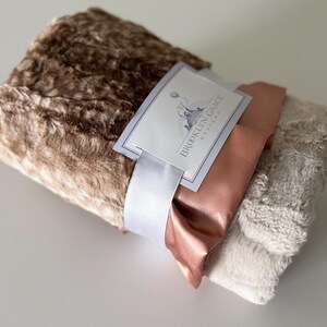 May include: A brown and beige faux fur blanket with a satin ribbon and a tag that reads "Brooklyn Grace Designs".