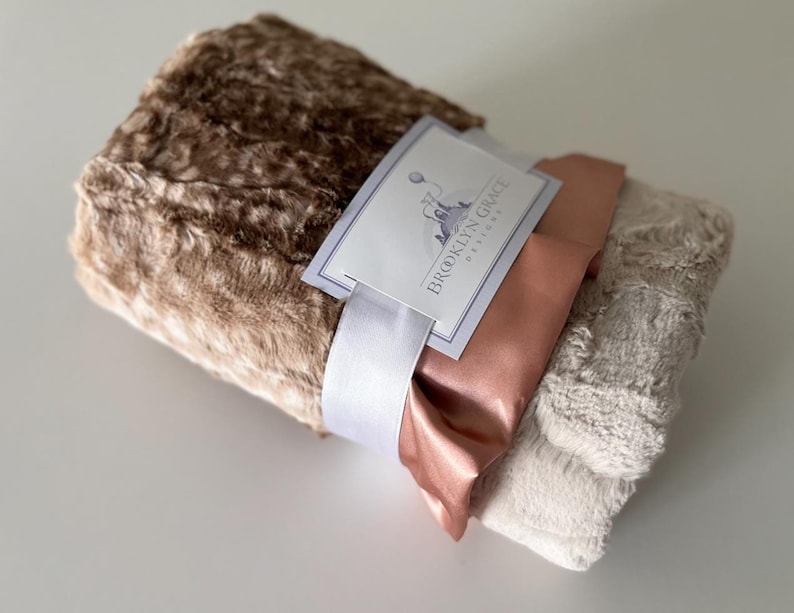 May include: A brown and beige faux fur blanket with a satin ribbon and a tag that reads "Brooklyn Grace Designs".