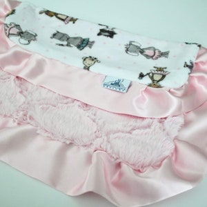 May include: A pink satin baby blanket with a ruffled edge and a soft, fluffy pink side. The blanket features a white side with a pattern of dancing animals in grey and brown.