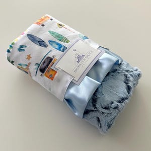 May include: A baby blanket with a white top layer featuring watercolor illustrations of surfboards, a camper van, and other beach-themed elements. The blanket has a blue satin trim and a soft, textured blue underside. A tag reads "Brooklyn Grace Designs."