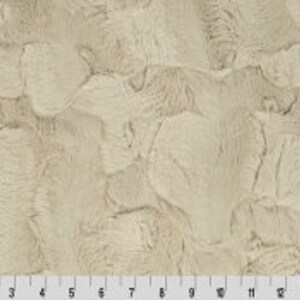 May include: A close-up of a beige faux fur fabric with a textured, wavy pattern. The fabric has a soft, plush feel.