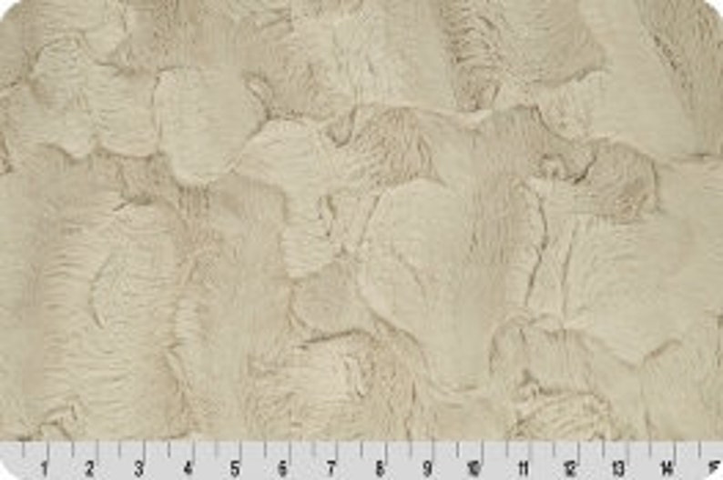 May include: A close-up of a beige faux fur fabric with a textured, wavy pattern. The fabric has a soft, plush feel.