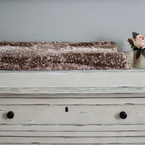 May include: A white changing table with a brown faux fur changing pad and a small vase with pink flowers on top.