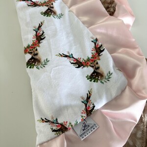 May include: A white baby blanket with a pink satin trim and a floral deer print. The blanket has a tag that says "Brooklyn Grace Designs".
