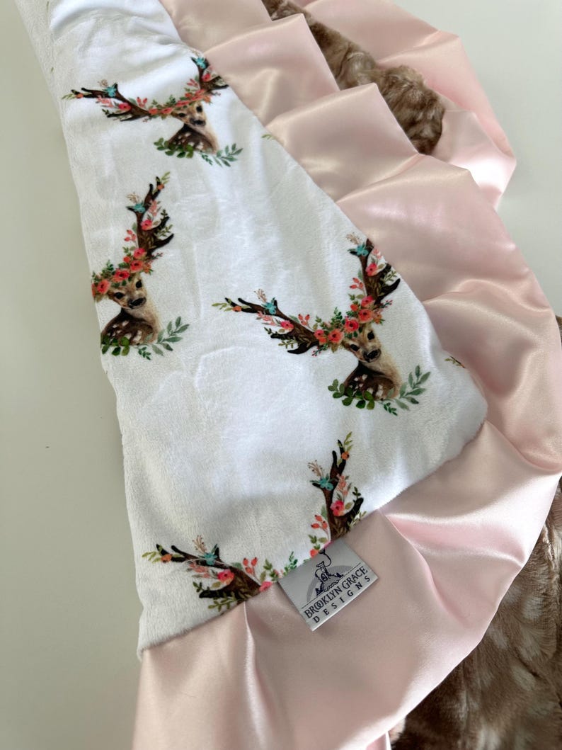 May include: A white baby blanket with a pink satin trim and a floral deer print. The blanket has a tag that says "Brooklyn Grace Designs".