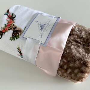 May include: A white and brown baby blanket with a floral deer print. The blanket is wrapped in a pink satin ribbon and a white satin ribbon with a tag that says "Brooklyn Grace Designs".
