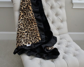 May include: A black and brown leopard print throw blanket with a black satin trim. The blanket is draped over a white tufted armchair.