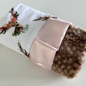 May include: A white baby blanket with a floral deer print on one side and a brown faux fur on the other side. The blanket has a pink satin trim.