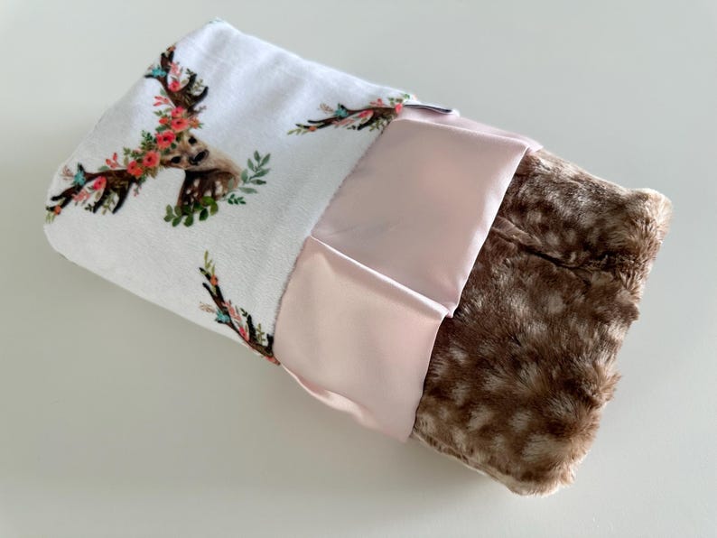 May include: A white baby blanket with a floral deer print on one side and a brown faux fur on the other side. The blanket has a pink satin trim.