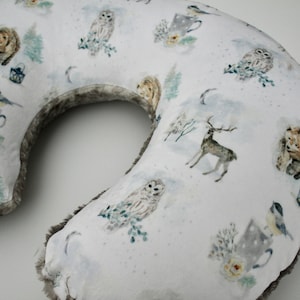 May include: A white and gray U-shaped pillow with a winter woodland print. The print features owls, deer, bears, birds, and trees. The pillow has a soft, fuzzy trim.
